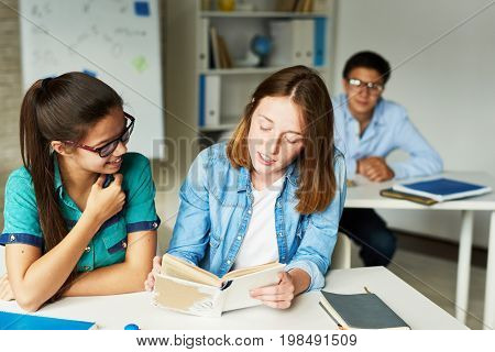 Portrait of two teenage girls reading book at desk together during lesson in modern classroom