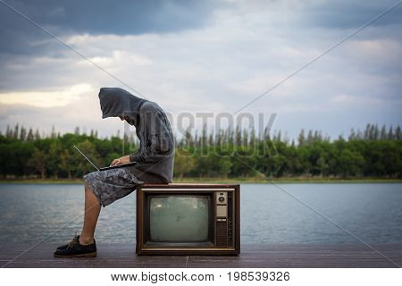 A Young Man Wearing A Hooded Sweatshirt Sitting Near The Old Television.a Hacker With A Laptop.