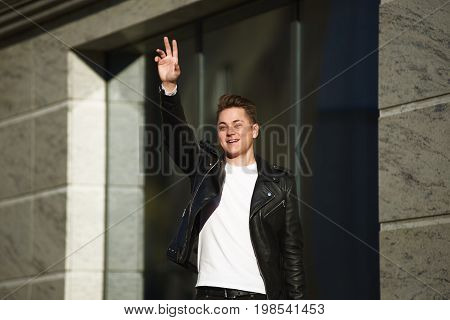 Fashionable smiling young European man wearing black leather jacket over white t-shirt standing outside modern buildin waving hand while noticing his friend across street trying to get attention