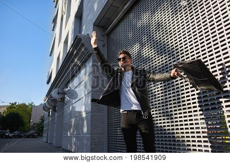 Outdoor lifestyle picture of handsome guy in shades standing on street holding bag in one hand and raising the other waiving it actively hailing a cab while being late to airport or urgent meeting