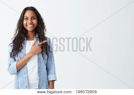 Fashionable emotional young African American female wearing denim shirt pointing her index finger at white blank wall background behind her, looking positive and happy, broadly smiling.