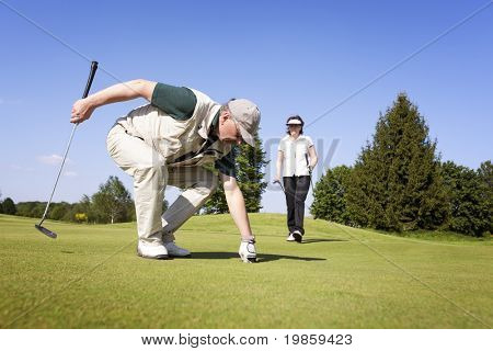 Senior actieve mannelijke golfspeler oppakken van een golfbal uit gat op groen met vrouwelijke golf speler walki