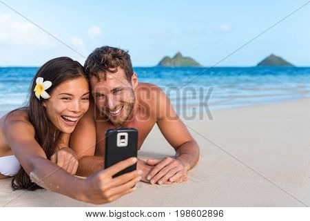 Beach vacation couple taking fun phone selfie on Hawaii vacation. Asian girl Caucasian man relaxing on Lanikai beach, Oahu, on summer holidays using smartphone for pictures together.