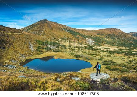 Hiker with backpack and trekking poles standing on a big stone near the mountain lake in a carpathian mountains