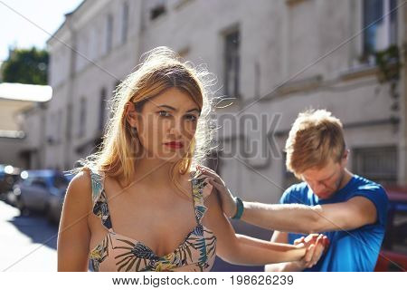 Desperate sad guy in blue t-shirt standing on street looking down and holding beautiful upset girl by the hand who is about to leave. Young couple having relationships problems has to break up