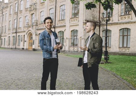 Two guys are standing together on the street near the university. They are talking about something and laughing. Apparently they are enjoying spending their time together.