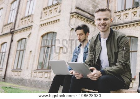 Two young guys are sitting on the bench near the university where they study. One of them is holding a laptop on his knees and reading some information from the laptop's screen. The other one of holding a tablet in his hands and smiling at the camera. Clo