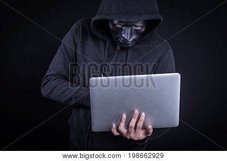 Anonymous male hacker using black mask to cover his face carrying laptop computer. Internet security and cyber attack concepts.