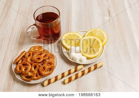 Transparent cup of black tea on a light table and saucers with pretzels sweet sticks and chopped lemon and pieces of refined sugar.
