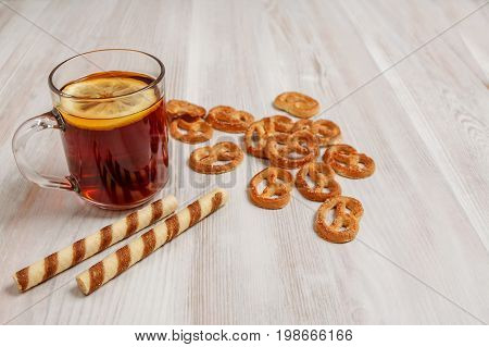 Transparent cup of black tea with lemon on a light table saucer with pretzels and sweet sticks.