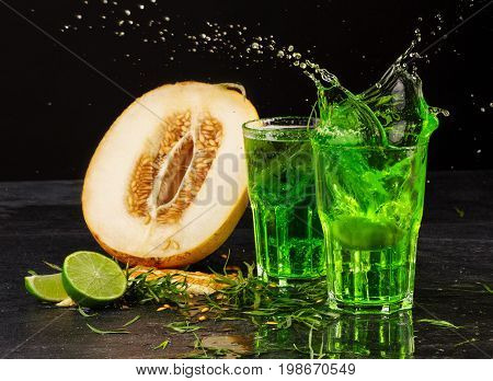 Close-up of two big glasses of green alcoholic drink with ice cubes and tarragon leaves on a black background. Refreshing summer beverages with ripe cut melon. Fresh and summer cold cocktails.