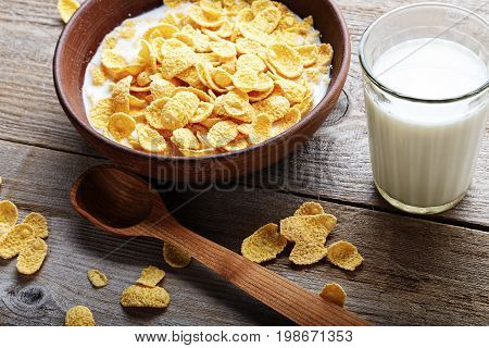 Cornflakes in a brown clay plate on a worn wooden background next is a glass of milk and a spoon and several flakes are scattered