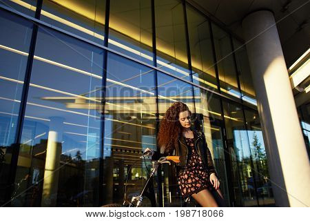 Urban summer shot of attractive young brunette Caucasian woman wearing floral pattern dress and leather jacket standing outside mall leaning on her vintage bicycle. People transportation and leisure