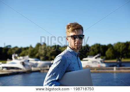 Staying connected. Outdoor shot of confident stylish young man with stubble wearing shades and shirt carrying generic laptop computer under his arm while spending weekend outdoors by the river
