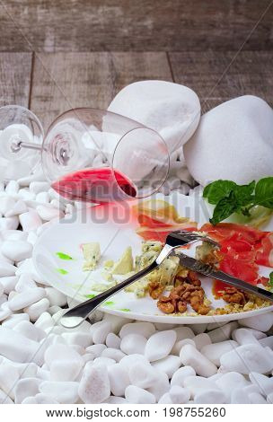 A beautiful composition of a delicatessen plate and a wineglass on white stones. A ceramic round plate with delicious snacks and a glass of semisweet red wine on a wooden background. Copy space.