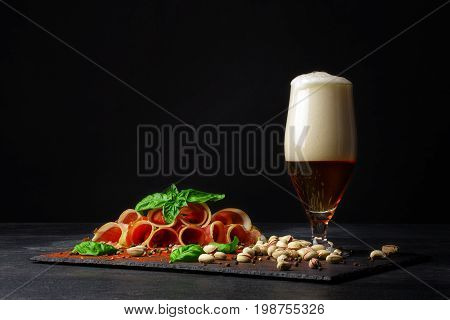 A beautiful composition of different snacks and a pint of saturated brown beer with thick froth on a black background. Pistachios and a plate of perfectly sliced balyk pieces. Copy space.