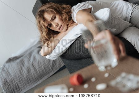 Being thirsty. Cheerless gloomy pretty woman stretching out her hand and taking a glass of water while being thirsty