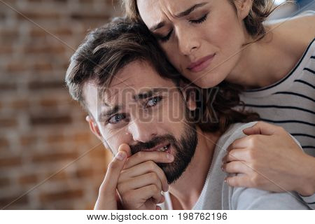 I need support. Unhappy crying young man sitting at the table and being hugged by his girlfriend while needing her support