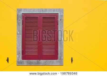 Bright yellow facade with a stone framed grey window covered with red shutter.