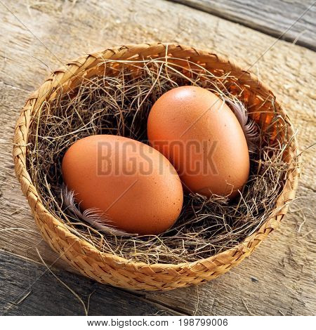 Couple of fresh chicken eggs in a rural basket with hey and feather on a wooden background. Close-up shot. Top view.
