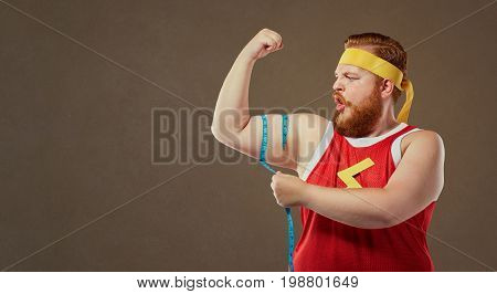 A funny fat freak man in sports clothes measures his arm with a centimeter.