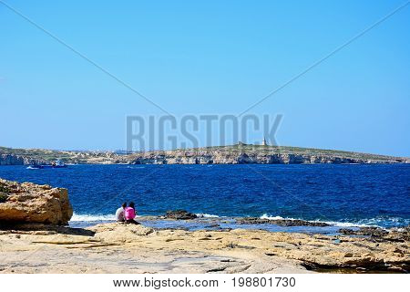 BUGIBBA, MALTA - MARCH 28, 2017 - A couple sitting on the rocky shoreline with views across the bay Bugibba Malta Europe, March 28, 2017.