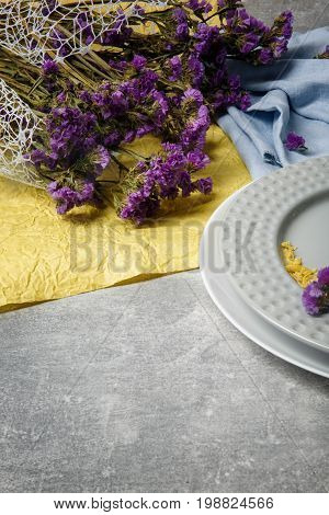 A colorful floral composition. A bouquet of little purple flowers on a blue and yellow fabric. Fancy plates on a gray table background. Health, relaxation and romance concept.