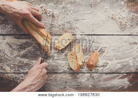 Baking background. Hands of baker cutting bread loaf with knife on rustic wooden table sprinkled with flour.