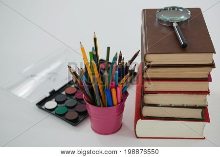 Close-up of book stack, magnifying glass, color pencils and palette on white background