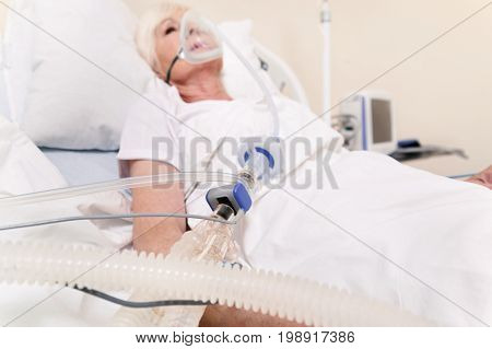 Vital mask. Feeble sore aged lady lying in bed while getting special treatment and wearing medical device for proper breathing