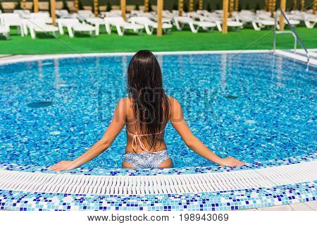 Beautiful Girl In A Bathing Suit Sits On The Edge Of The Pool, The View From The Back