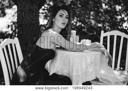 Young Woman In A Long Evening Dress Sits At A Table In The Woods. Black And White Photography