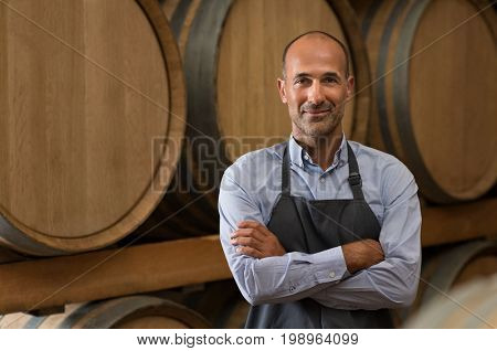 Satisfied winemaker in cellar with folded hand. Happy man standing in his warehouse and looking at camera. Smiling professional man standing in wine cellar with wine wooden barrels in background.