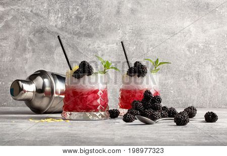 Two glasses with fruit cocktails, black drinking straws, fresh blackberries, green sappy leaves of mint and silver metallic shaker on a grey light background.
