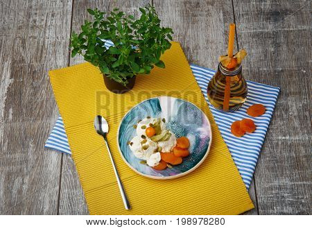 Top view of white sweet ice-cream with dried orange apricots, fresh juicy physalis, pumpkin seads, shining metallic tea spoon, little green tree, yellow and striped napkins , bottle with beverage on a wooden background.