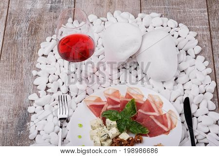 Top view of a plate with slices of prosciutto seasoned with leaves of bazil, cubes of Roquefort cheese and walnuts, silver fork and knife on a white stone wooden background, little white stones. Expensive food.