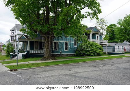 CADILLAC, MICHIGAN / UNITED STATES - MAY 31, 2017: A large elegant blue Victorian mansion, with a wraparound porch, a balcony, and a maple shade tree, in Cadillac's Courthouse Hill Historic District.
