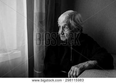 An elderly woman in his house looks out the window longingly. Old lady retired. Black and white photo.