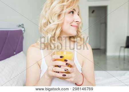 Attractive Blonde Woman Sits On The Edge Of The Bed At Home And Holds A Stocker With Orange Juice In