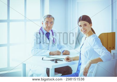 A doctor smiling at the camera with her male colleage in the back of the consulting room in hospital