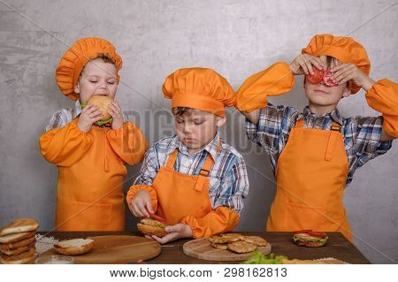 Three Cute Boys In Costumes Cooks Engaged In Cooking Homemade Burgers. Three Brothers Preparing Fami