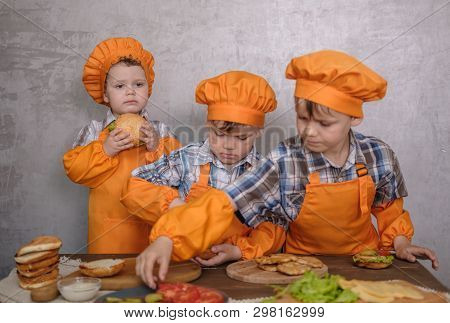Three Cute Boys In Costumes Cooks Engaged In Cooking Homemade Burgers. Three Brothers Preparing Fami