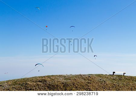 Paraglider On Blue Sky With Empty Bench On Foreground. Borso Del Grappa, Famous Paragliding Site, It