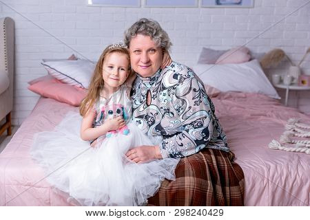 Happy Little Girl And Her Grandmother Are Sitting Together And Hugging In The Bedroom. They Are Smil