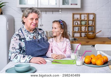 Happy Little Girl And Her Grandmother Have Breakfast Together In A White Kitchen. They Are Having Fu
