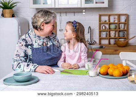 Happy Little Girl And Her Grandmother Have Breakfast Together In A White Kitchen. They Are Having Fu