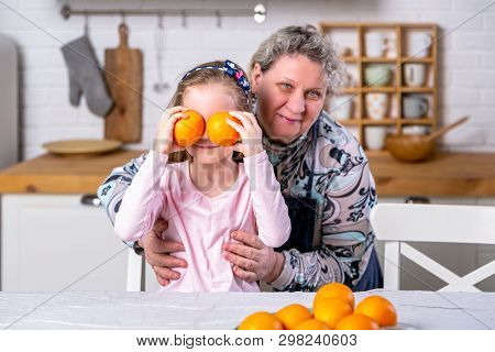 Happy Little Girl And Her Grandmother Have Breakfast Together In A White Kitchen. They Are Having Fu
