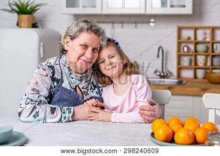 Happy Little Girl And Her Grandmother Have Breakfast Together In A White Kitchen. They Are Having Fu