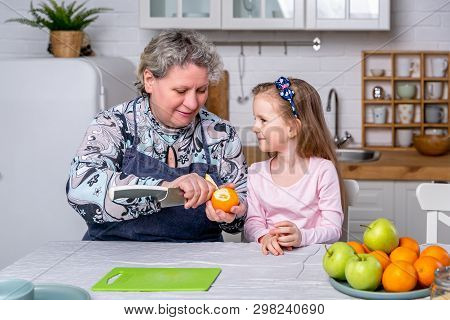 Happy Little Girl And Her Grandmother Have Breakfast Together In A White Kitchen. They Are Having Fu