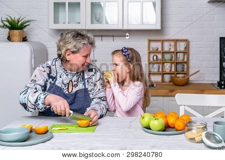Happy Little Girl And Her Grandmother Have Breakfast Together In A White Kitchen. They Are Having Fu